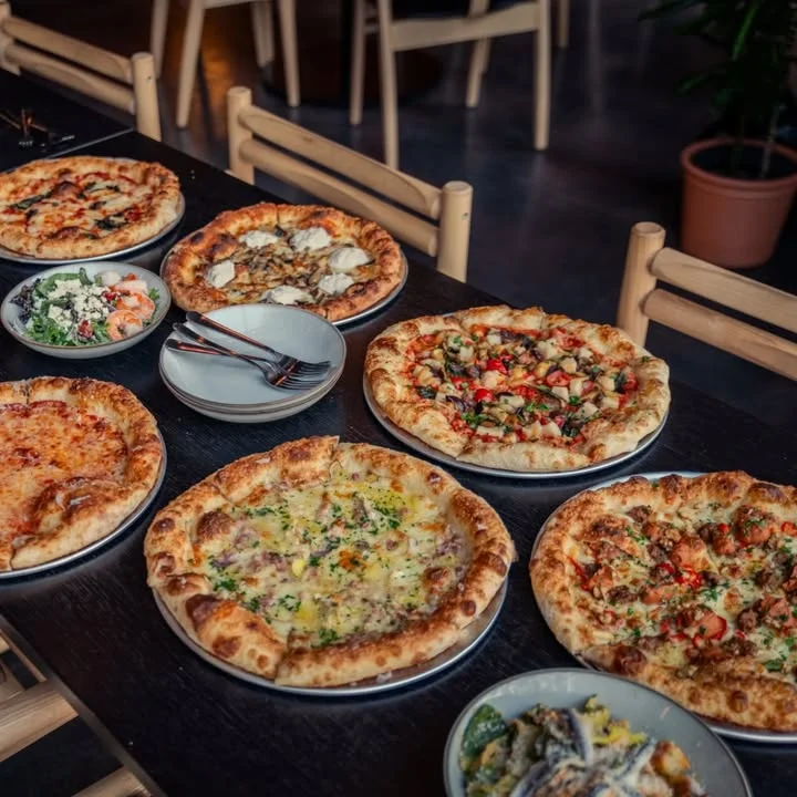 A variety of pizzas displayed on a table, alongside salads and utensils. Each pizza has different toppings.