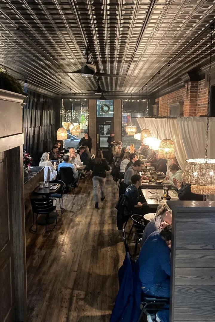 An elegant restaurant interior featuring wooden floors, large windows, and woven pendant lighting. The tables are neatly arranged for dining.
