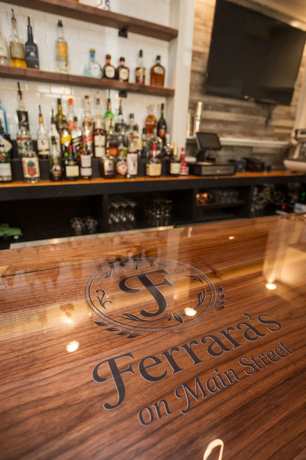 A bar top with a polished wooden surface displaying the logo of Ferrara's on Main Street. Behind the bar, various bottles of spirits are organized on shelves.