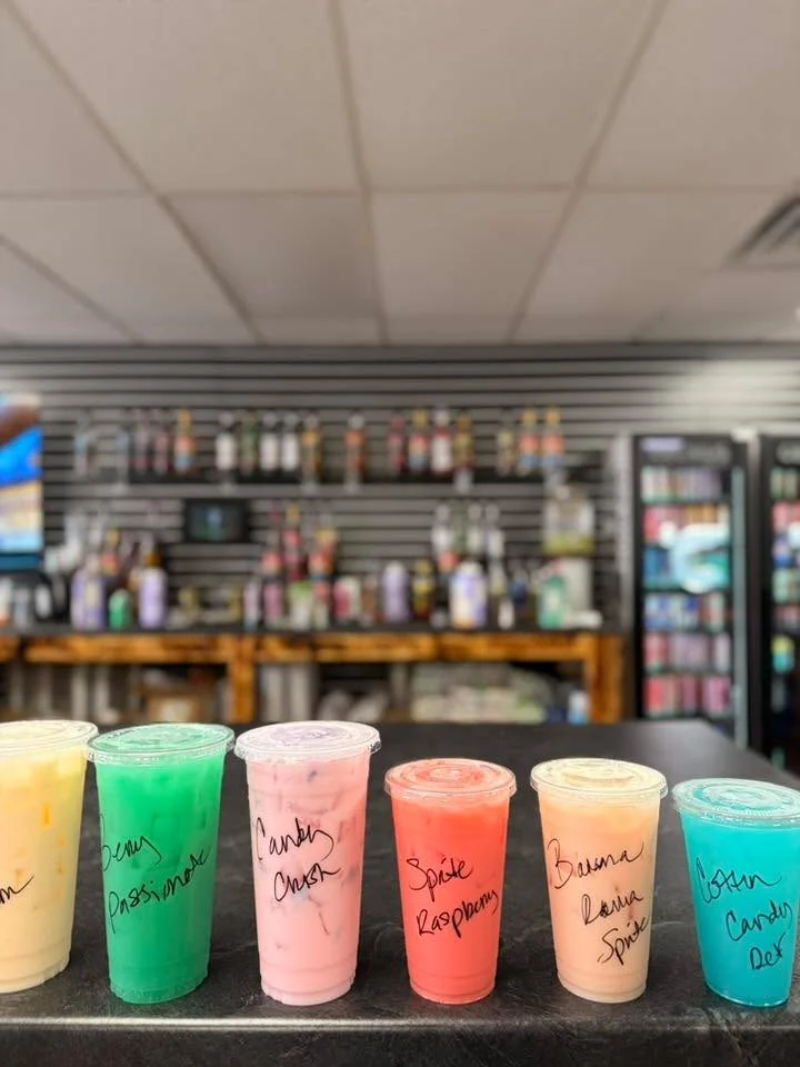 A lineup of six colorful drinks in clear cups, labeled with different names, set on a counter in a store.