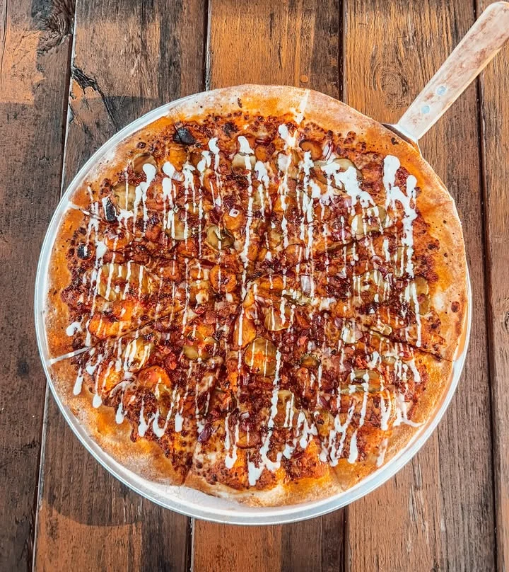 A round pizza topped with various ingredients including pickles and drizzled with a sauce on a wooden table.