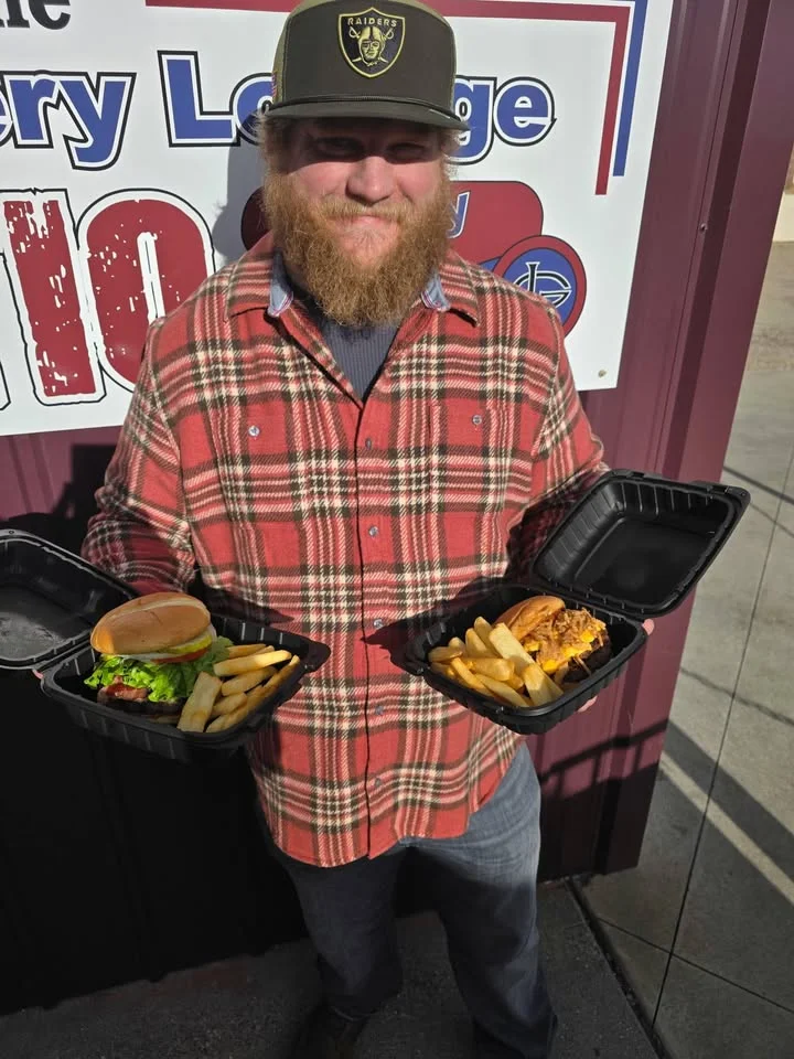 A person holding two takeout containers, featuring a burger with toppings and fries, standing outside a food establishment.