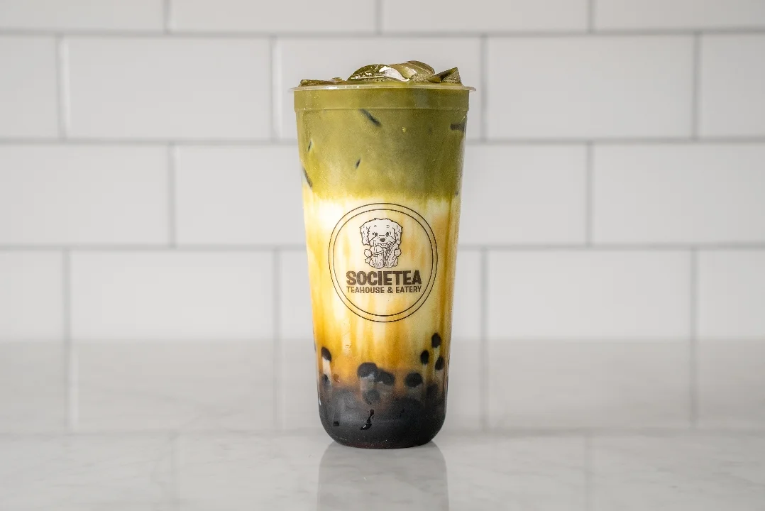 A colorful layered beverage featuring green tea, ice, and boba at the bottom. The drink is served in a clear cup with a logo.