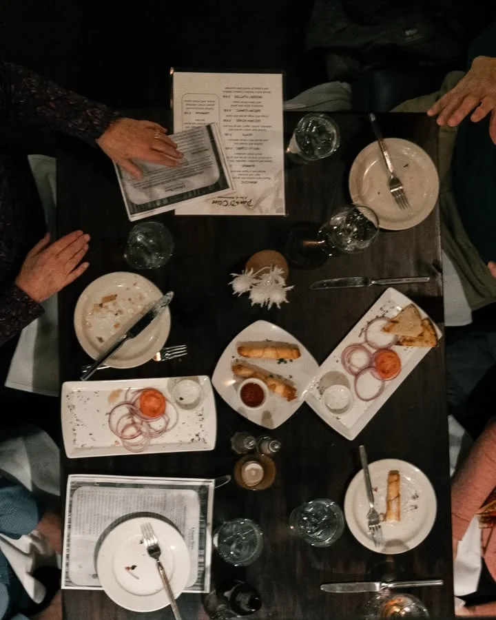 An overhead view of a dining table with plates of food, utensils, and menus, surrounded by dining guests.