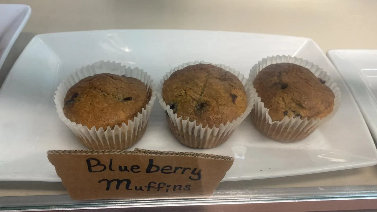 Three blueberry muffins in white liners, arranged on a white plate with a small sign identifying them.