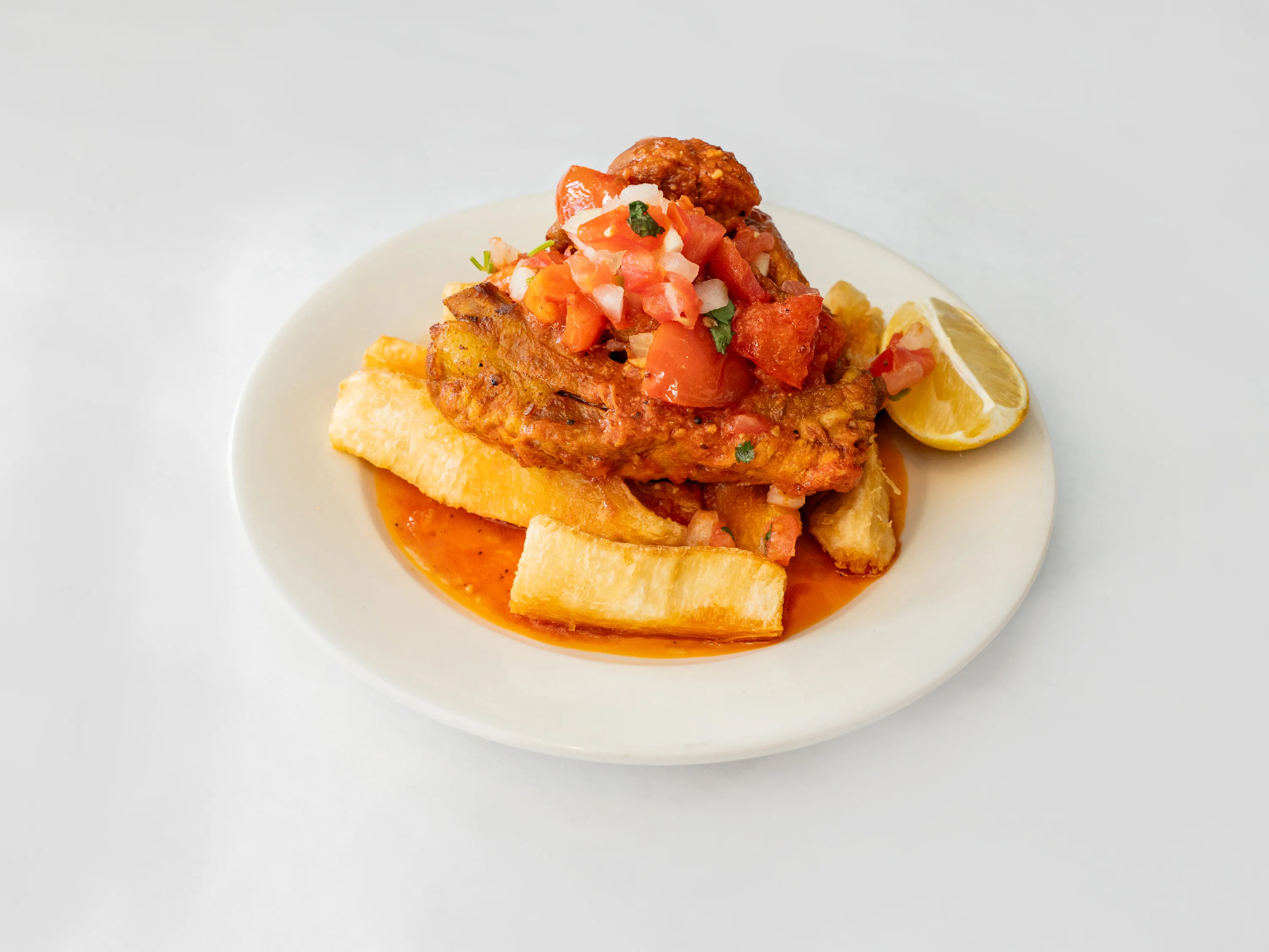 A plate featuring fried fish topped with fresh tomato salsa and served over cassava. A slice of lime accompanies the dish.