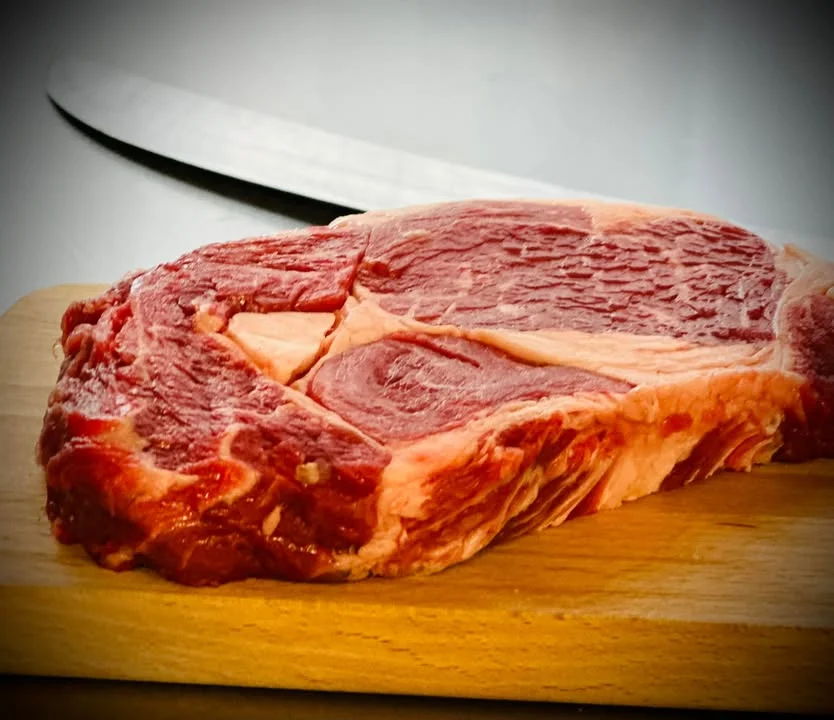 A raw ribeye steak placed on a wooden cutting board, showcasing its marbled texture, with a knife visible in the background.