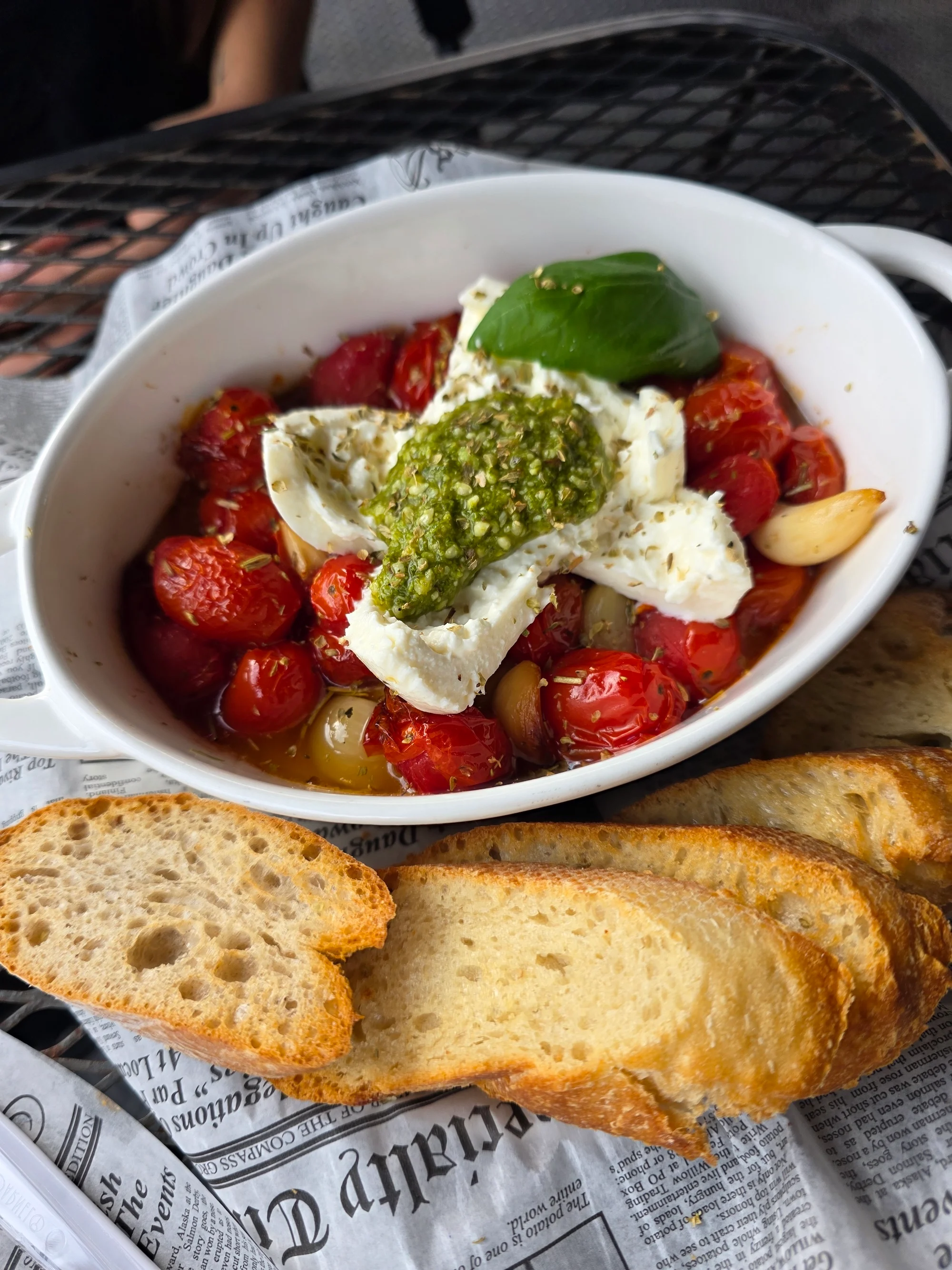 A delicious dish featuring baked cherry tomatoes, mozzarella, and pesto, served with slices of toasted bread on a table.