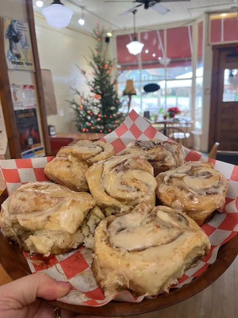 A hand holding a plate of cinnamon rolls drizzled with frosting, decorated with red and white checkered paper. A Christmas tree can be seen in the background.
