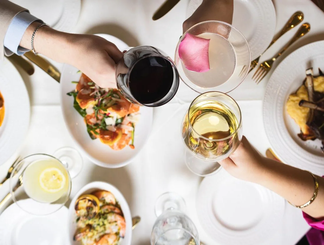 A beautifully plated dish of lamb chops garnished with herbs, surrounded by colorful vegetables and emitting wisps of steam, paired with a glass of red wine.