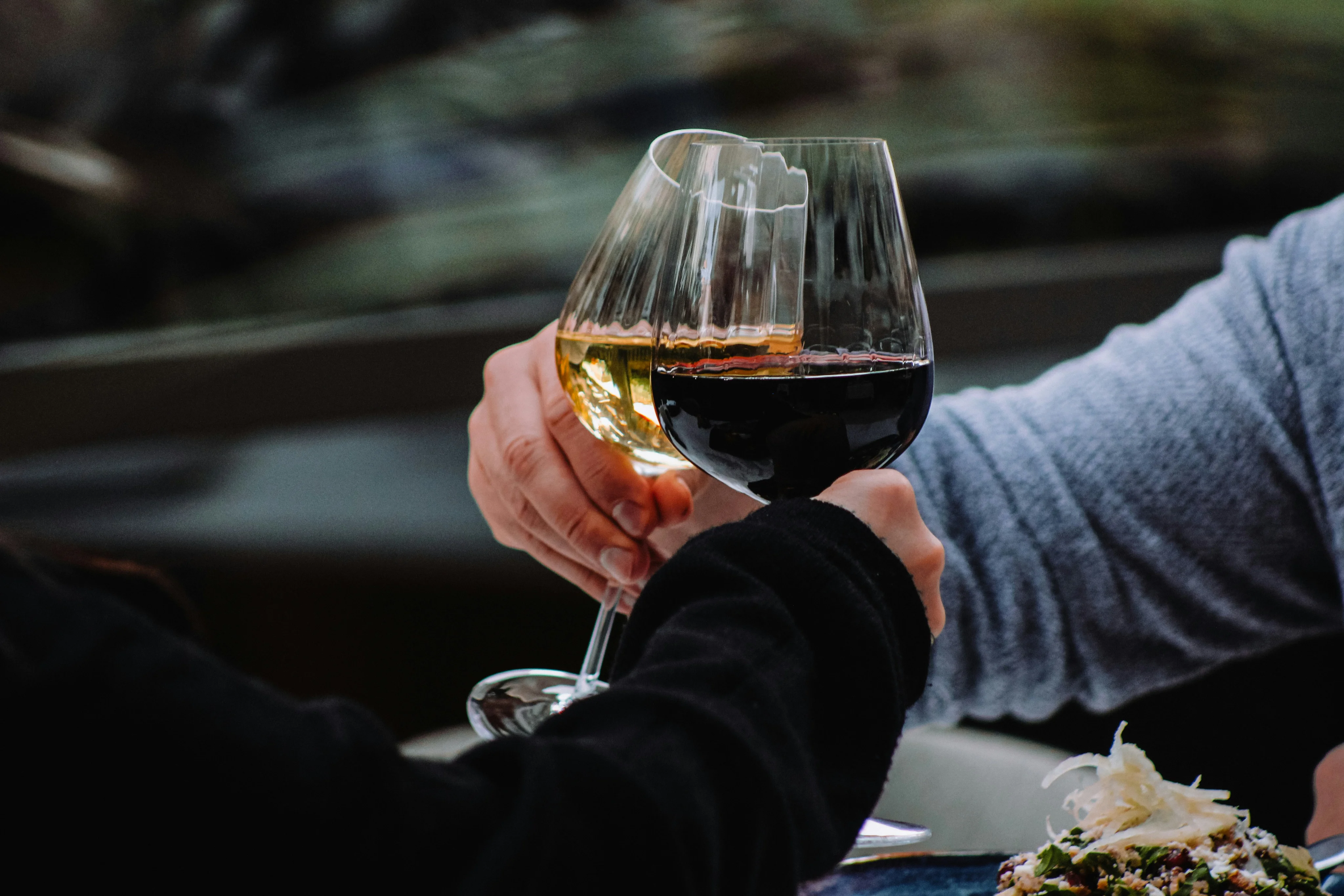 Two glasses clinking together in a toast, one filled with red wine and the other with white, set against a blurred background.