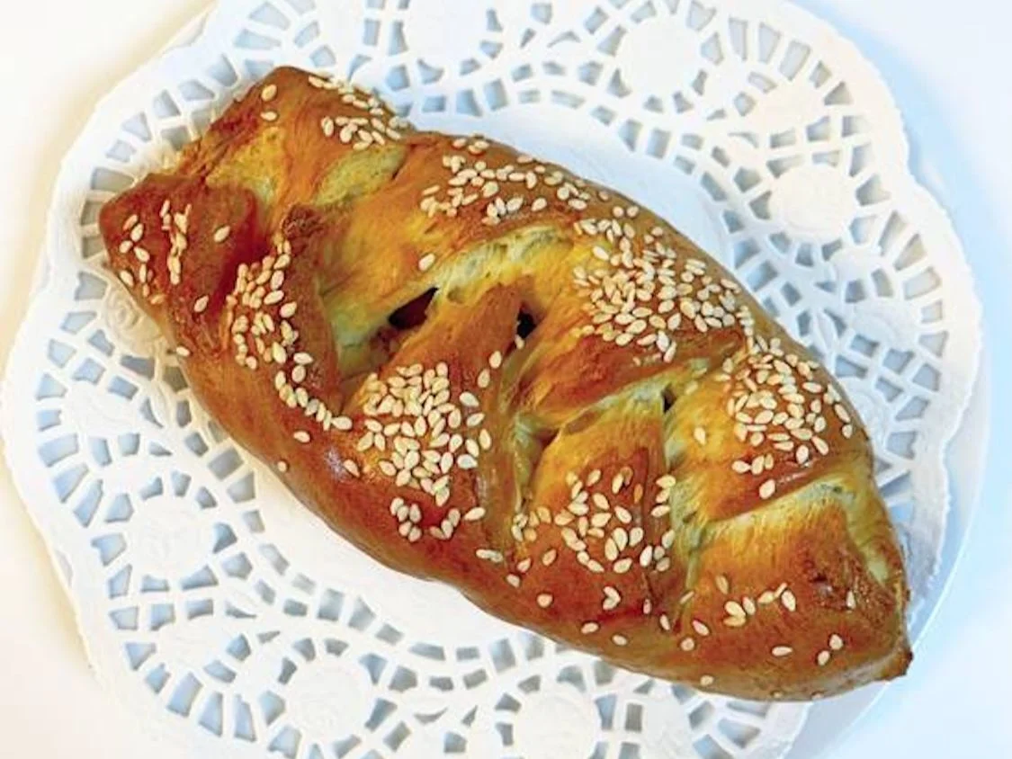 A braided pastry with a glossy finish, sprinkled with sesame seeds, served on a decorative doily.