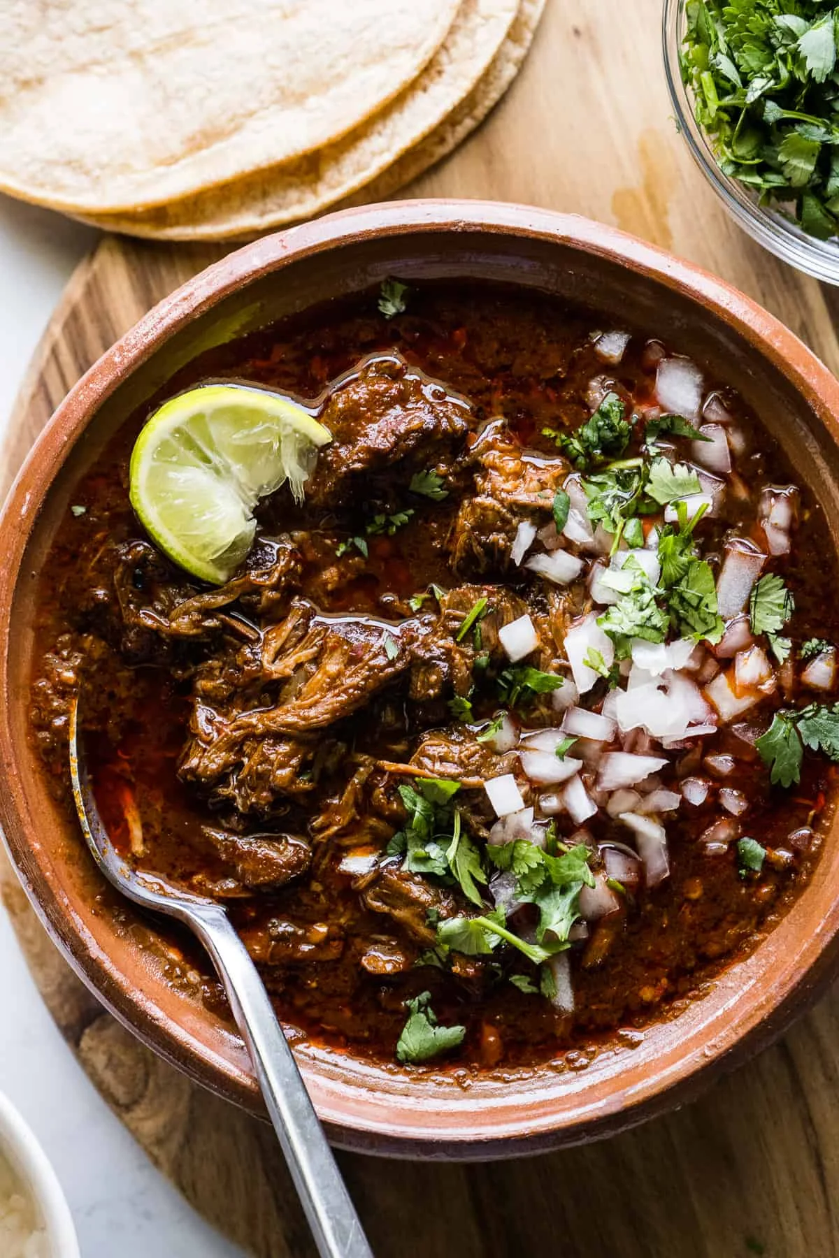 A hearty dish of slow-cooked meat in a rich, red sauce, garnished with chopped onions, cilantro, and a lime wedge, served with tortillas on the side.
