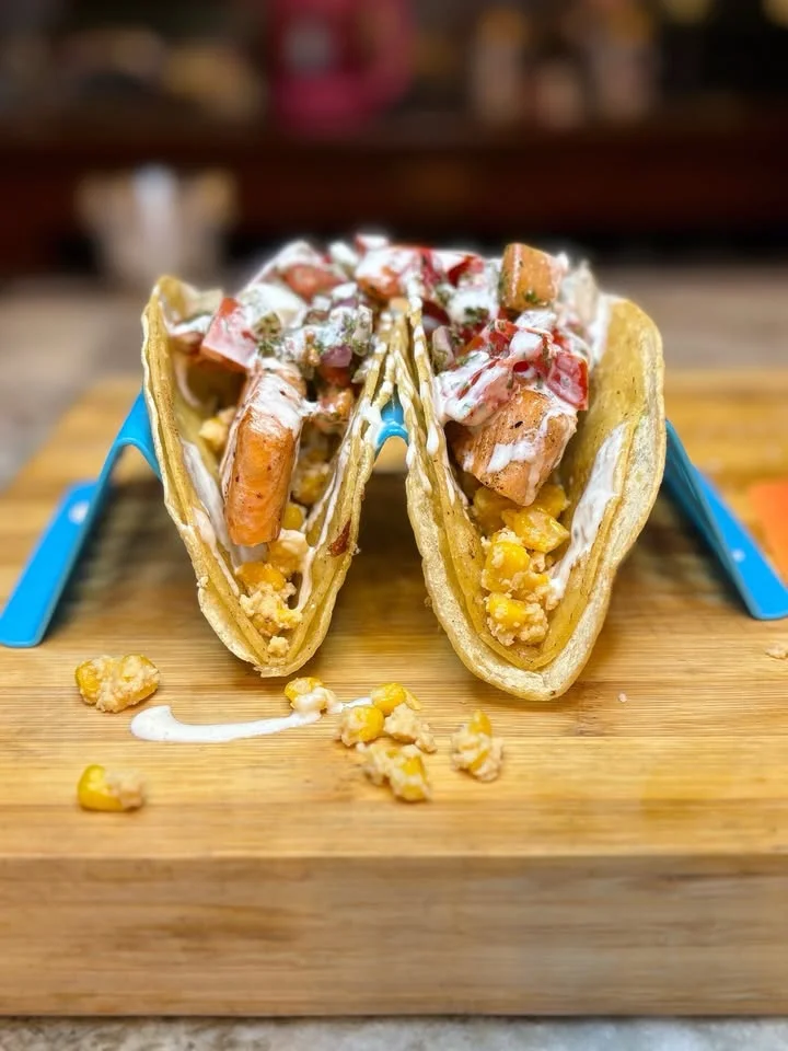 A pair of tacos filled with grilled meat, garnished with toppings such as onions, lettuce, and a creamy sauce, served on a wooden cutting board.