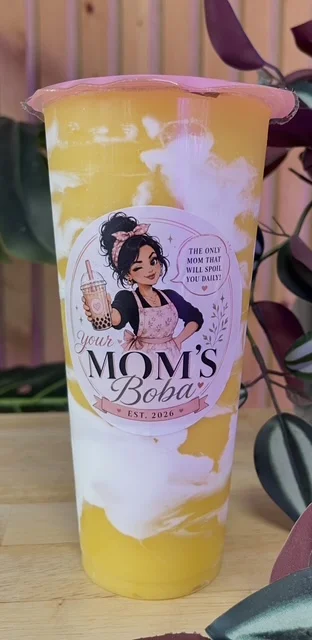 A transparent cup containing a layered yellow and white drink, topped with a colorful label that features an illustration and text related to boba.
