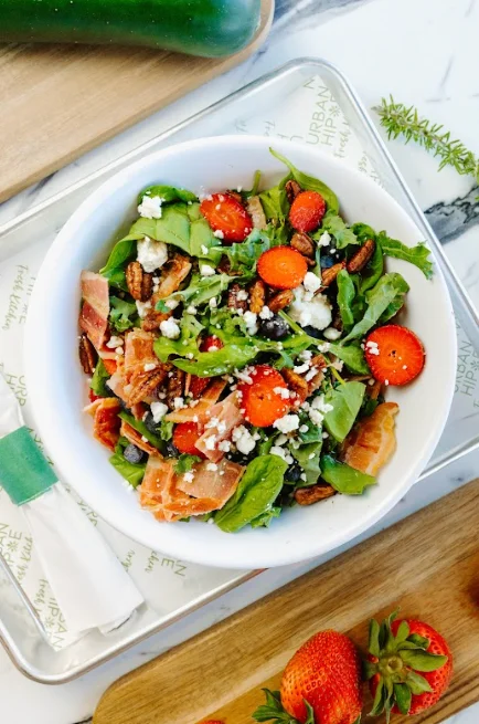 A fresh salad featuring mixed greens, strawberries, pecans, and cheese in a bowl, surrounded by colorful ingredients.
