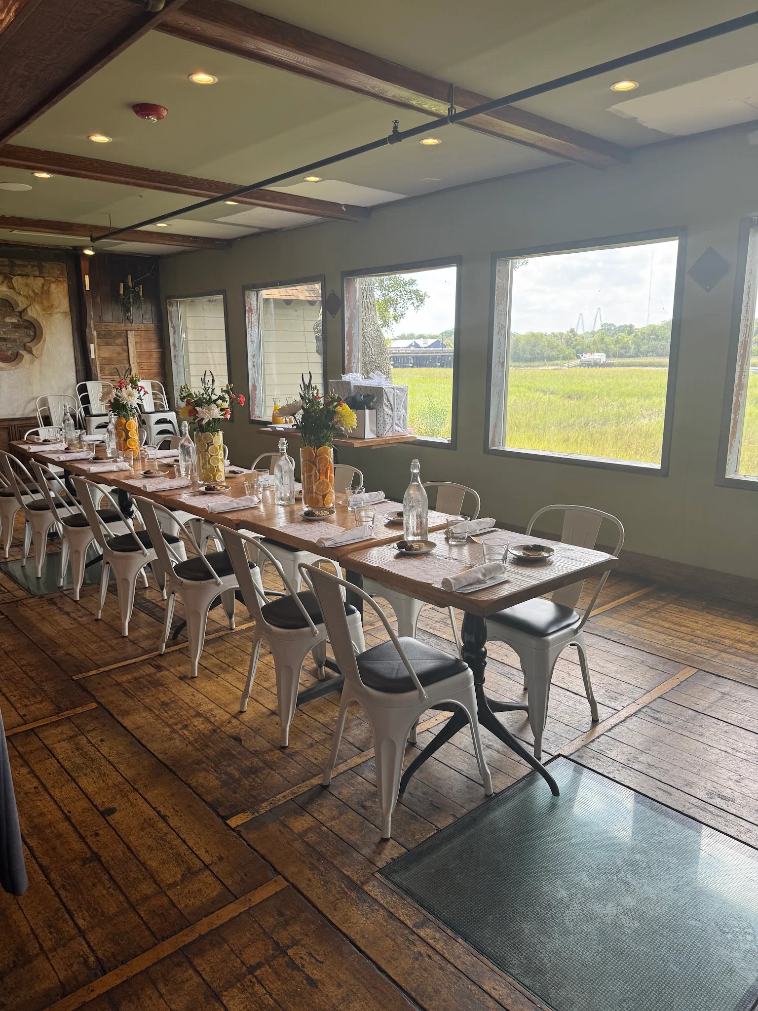 Long communal table set for dinner in the dining room of The Mill Street Tavern, with windows overlooking the Shem Creek marsh