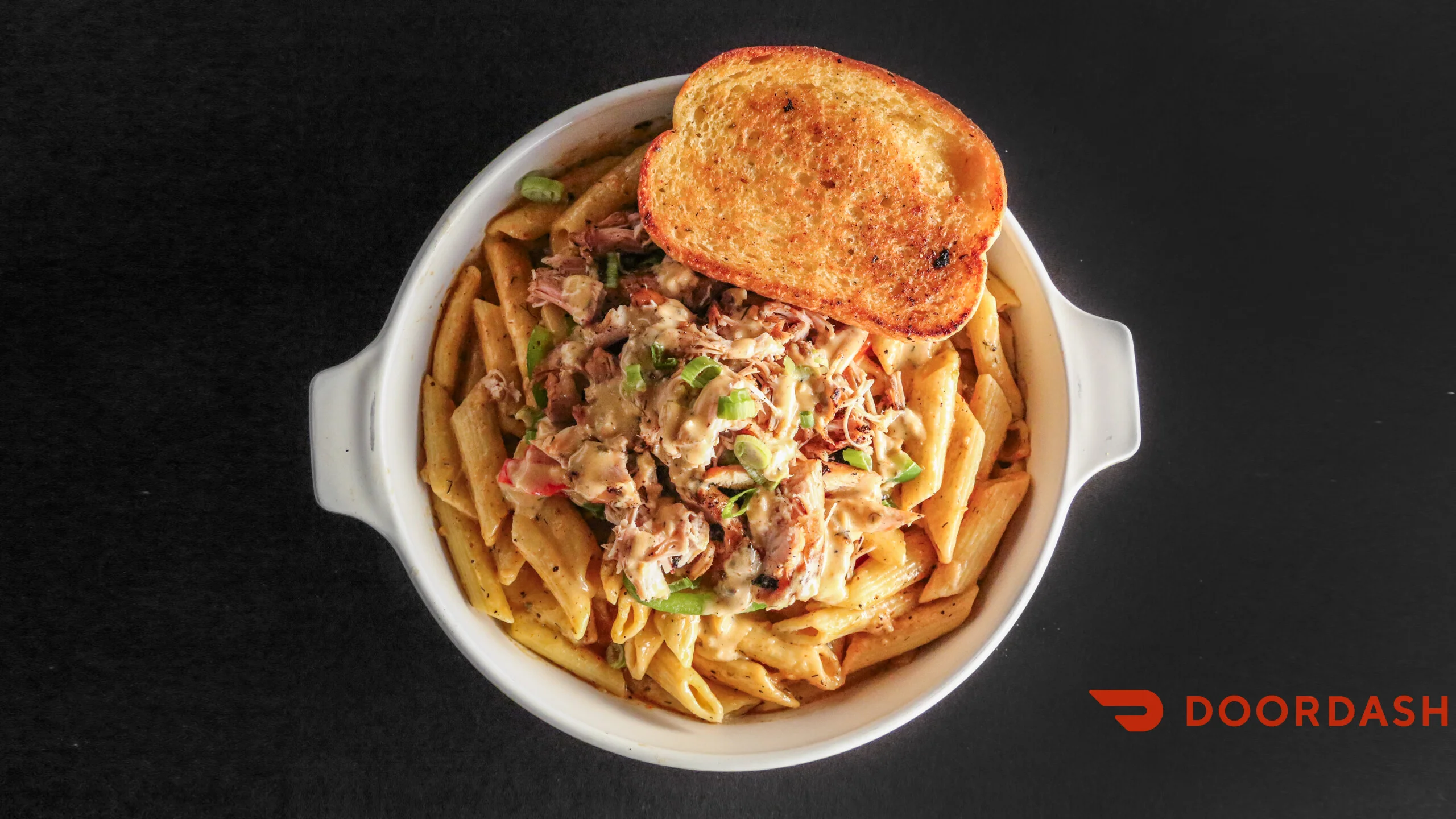 A bowl of pasta topped with shredded chicken and green peppers, garnished with slices of toasted bread. The dish features penne pasta in a creamy sauce.