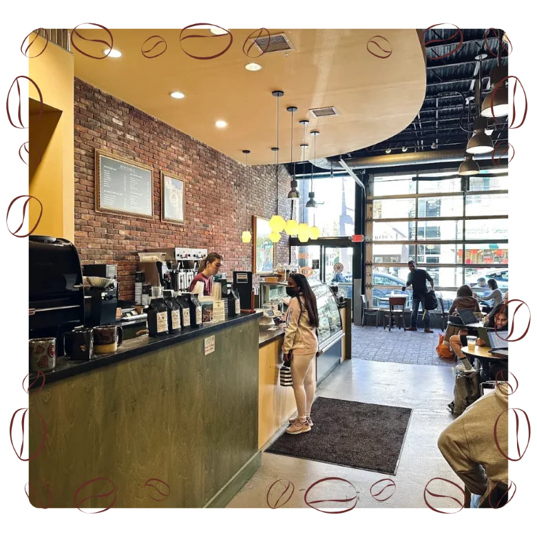 A cozy café with warm lighting and a brick wall. A customer orders at the counter, while others sit at tables using laptops, creating a busy, relaxed atmosphere.
