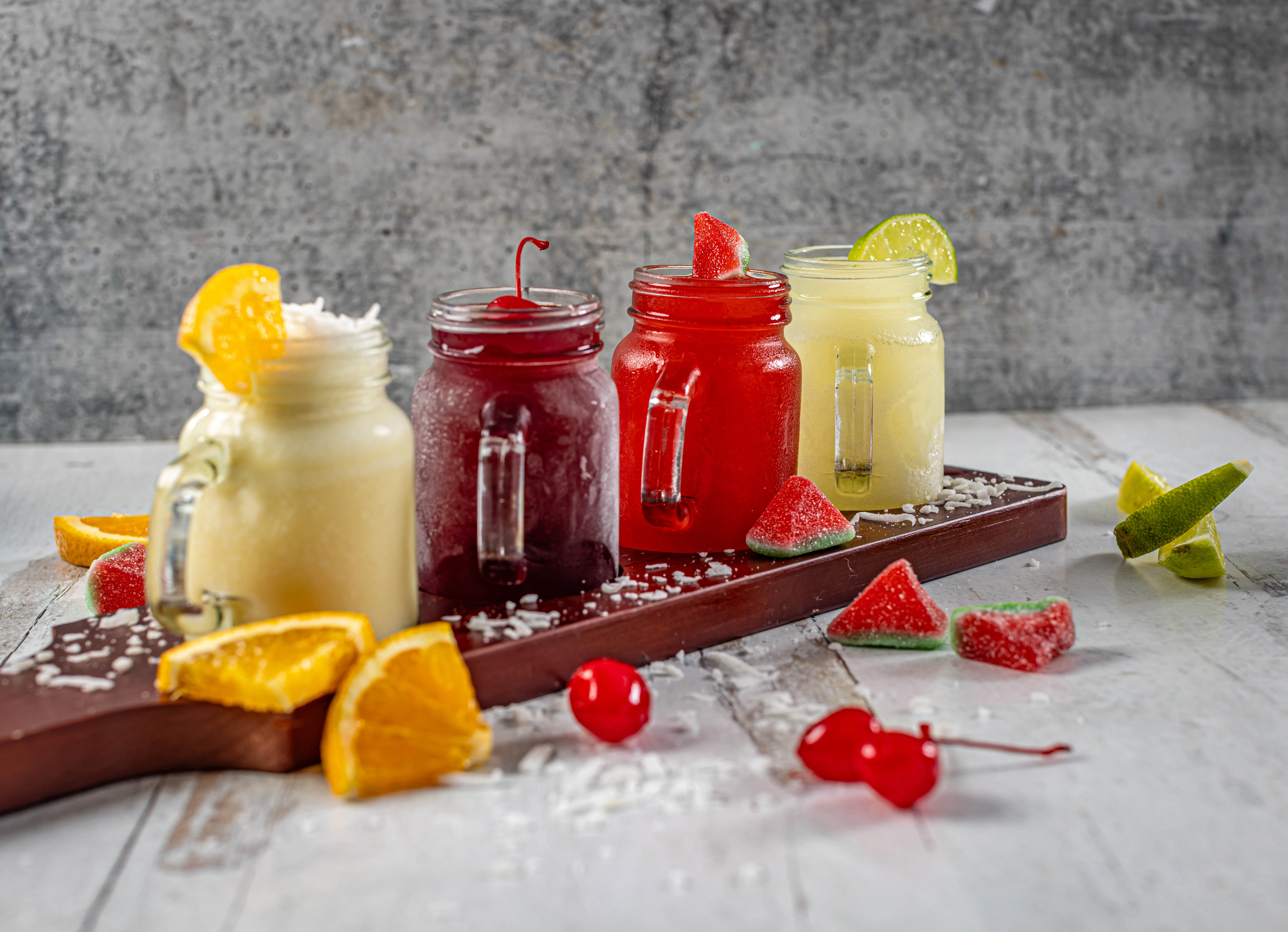 A colorful flight of four refreshing fruit juices, served in mason jar mugs and garnished with fresh fruit and candy.