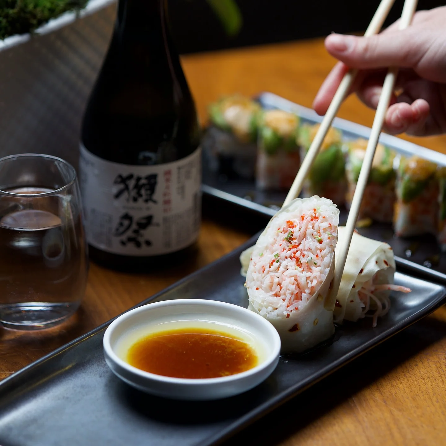 Fresh crab hand rolls served with a citrus dipping sauce, accompanied by a bottle of premium sake and a variety of specialty sushi rolls in the background.