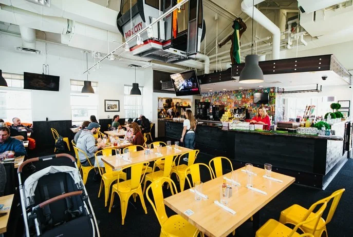 Another angle of a lively restaurant featuring wooden tables and bright yellow chairs. People are dining and enjoying their meals, with a bar area in the background.