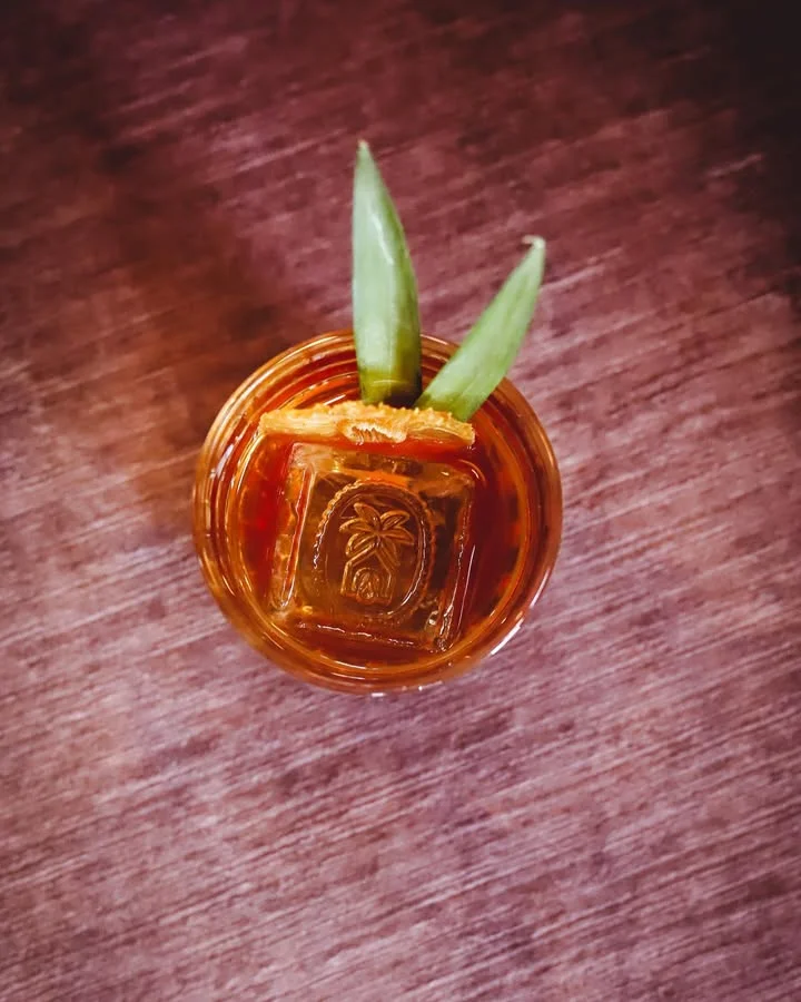 An overhead view of a cocktail garnished with green leaves and an ice cube embossed with a palm tree motif.
