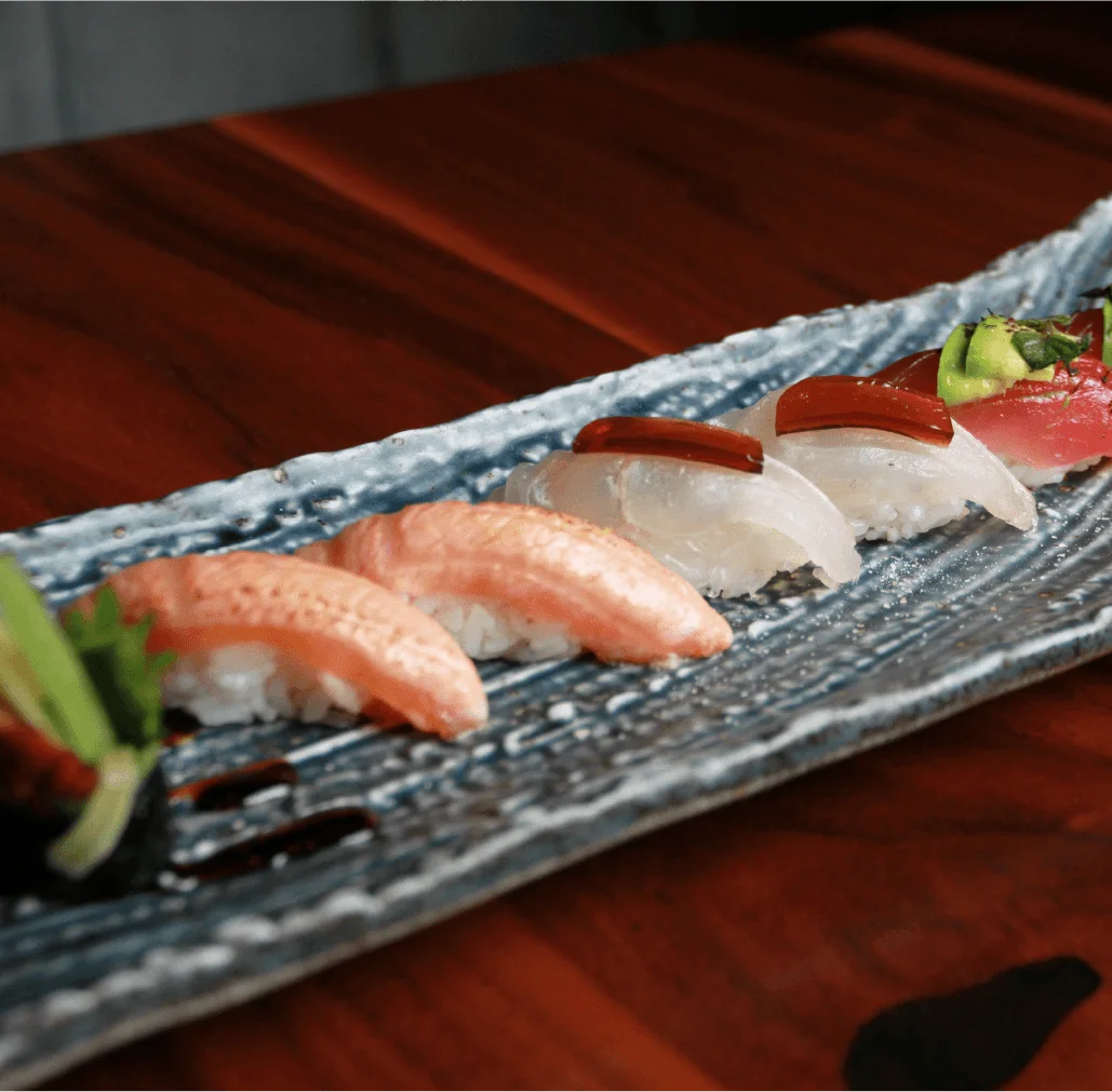 An assortment of pairs of nigiri and gunkan sushi, featuring tuna topped with avocado, white fish with a dark glaze, seared salmon, and eel rolls garnished with green scallions, beautifully arranged on a long, textured blue-grey ceramic plate over a wooden table surface.