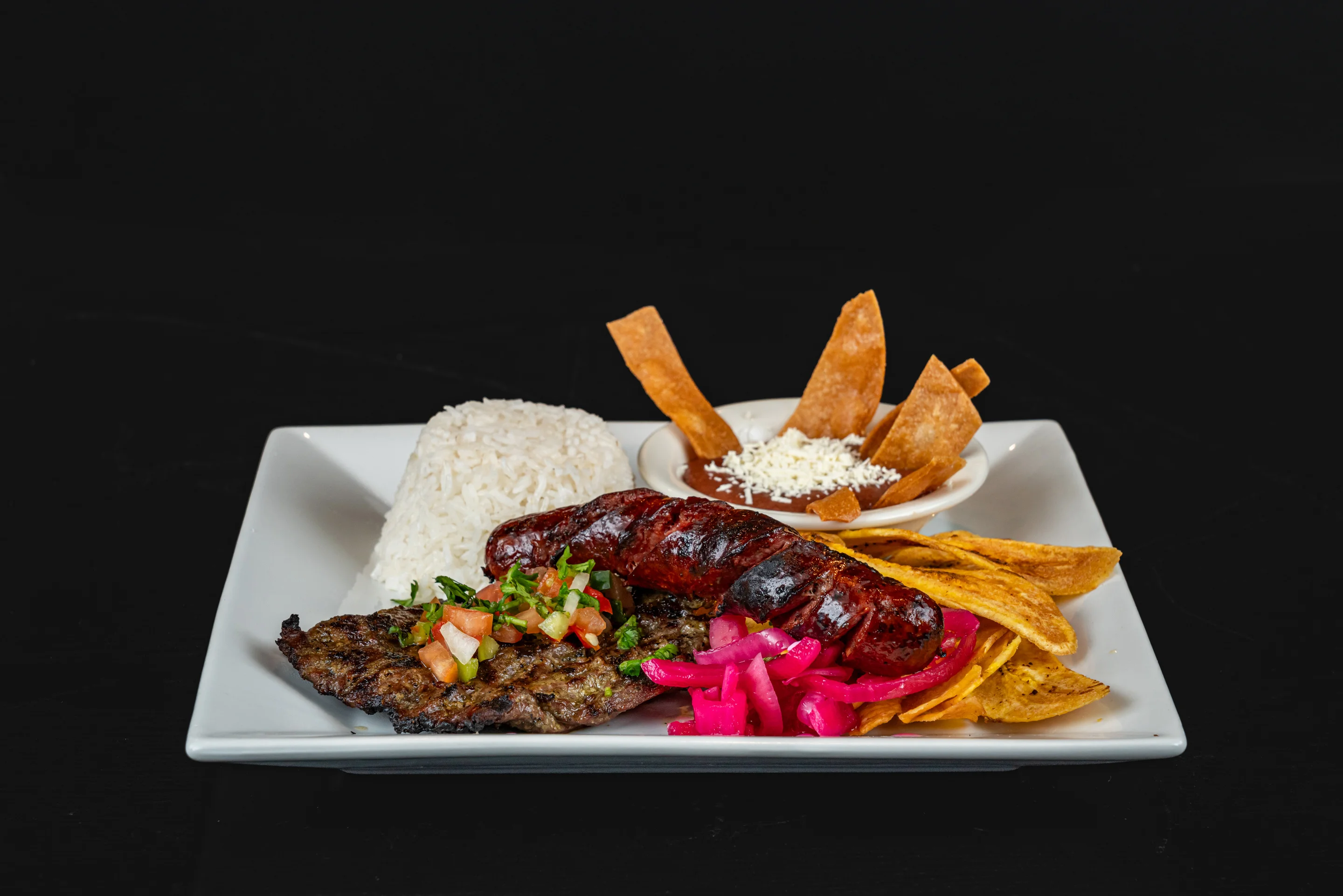 A hearty plate featuring grilled meat, chorizo, white rice, and colorful accompaniments like pickled onions and fresh salsa, alongside crispy tortilla chips and a rich dip.