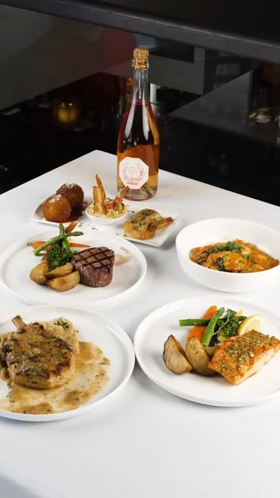 An assortment of gourmet dishes including steak, salmon, and a variety of sides, complemented by a bottle of sparkling wine.