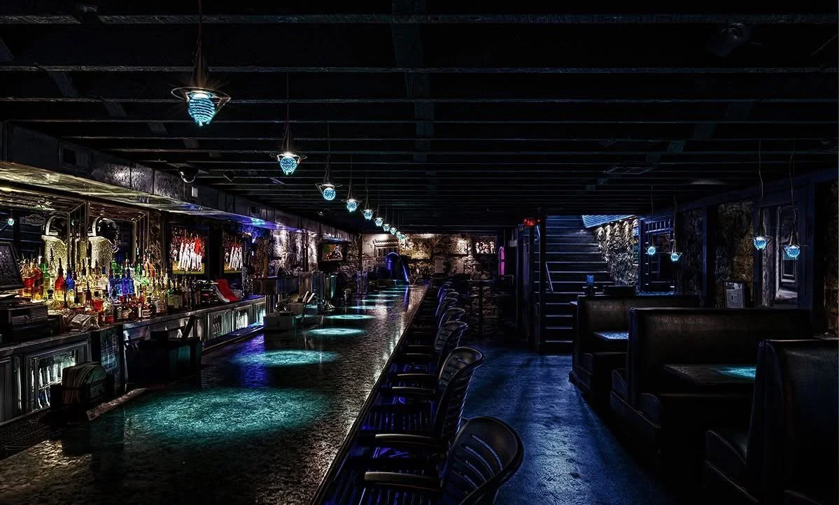 An illuminated bar area featuring a long counter, colorful drinks, and a stylish ambiance. The atmosphere is dimly lit with modern fixtures and a staircase in the background.