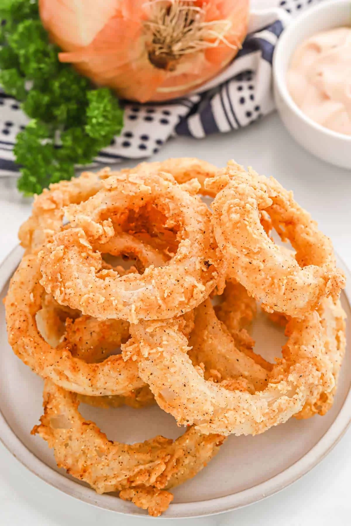 A plate piled with crispy onion rings, lightly seasoned and golden brown, accompanied by a background of fresh onion and greens.