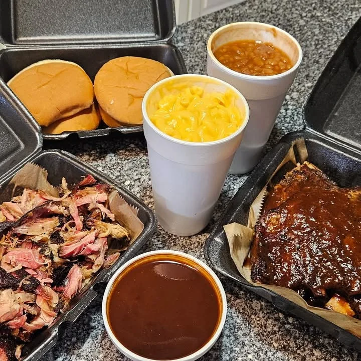VooDoo BBQ takeout meal featuring sauced ribs, pulled pork, mac and cheese, baked beans, buns, and mojo sauce in Pensacola.