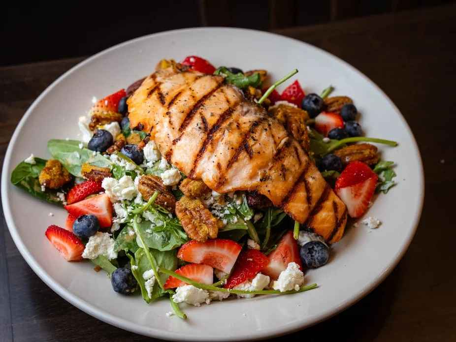 A fresh salad topped with grilled salmon, mixed greens, strawberries, blueberries, and feta cheese, presented on a round white plate.