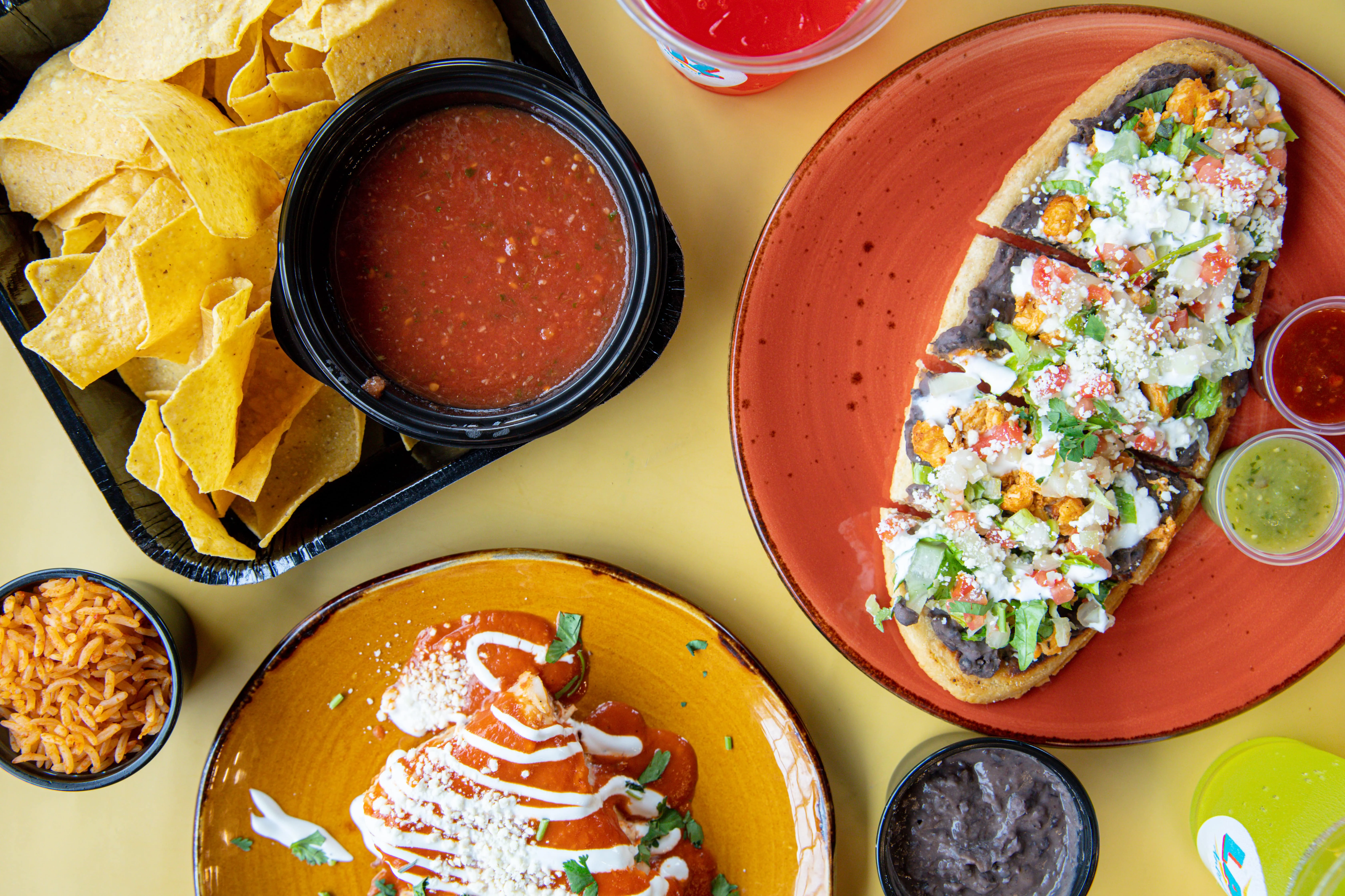 A colorful spread of Mexican cuisine featuring crispy tortilla chips, a bowl of salsa, a topped tortilla, rice, and black beans, beautifully arranged on a yellow background.