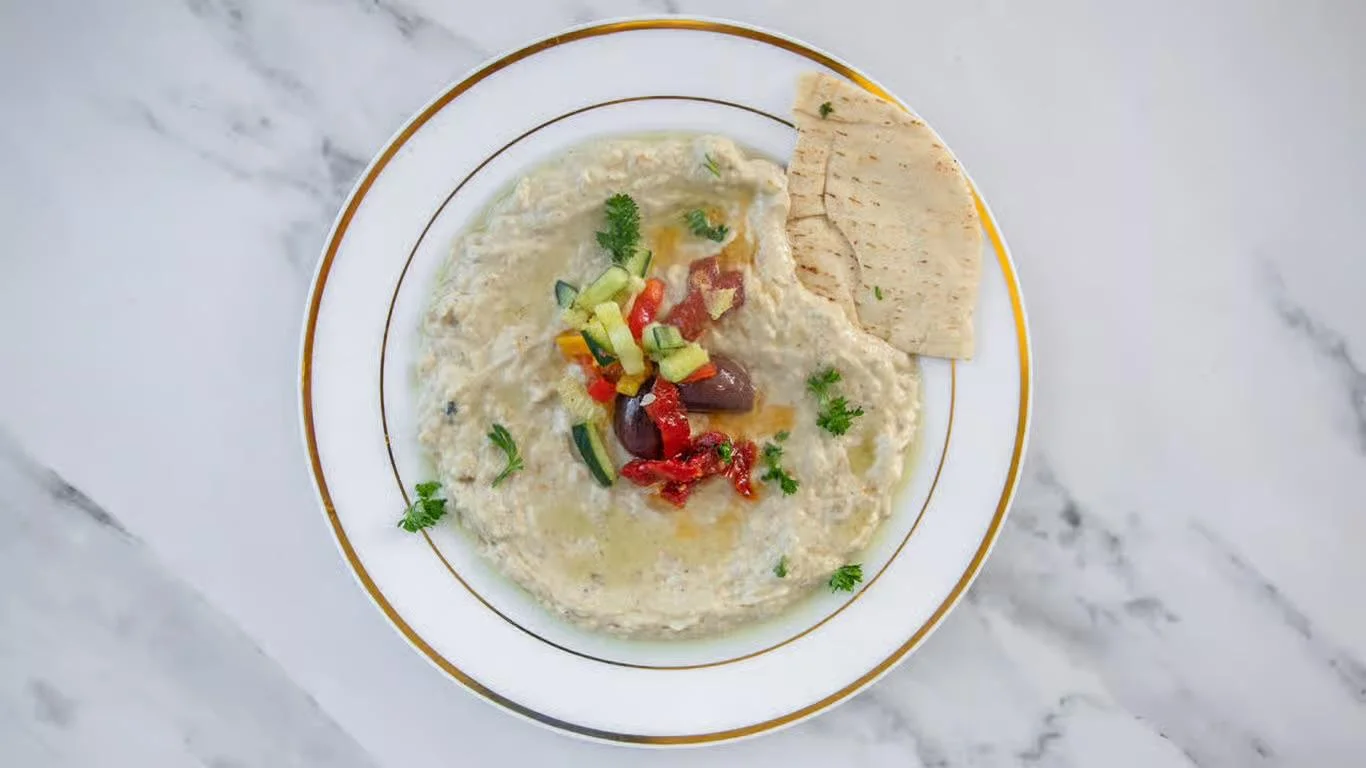 A serving of creamy hummus topped with fresh vegetables and surrounded by pieces of pita bread.