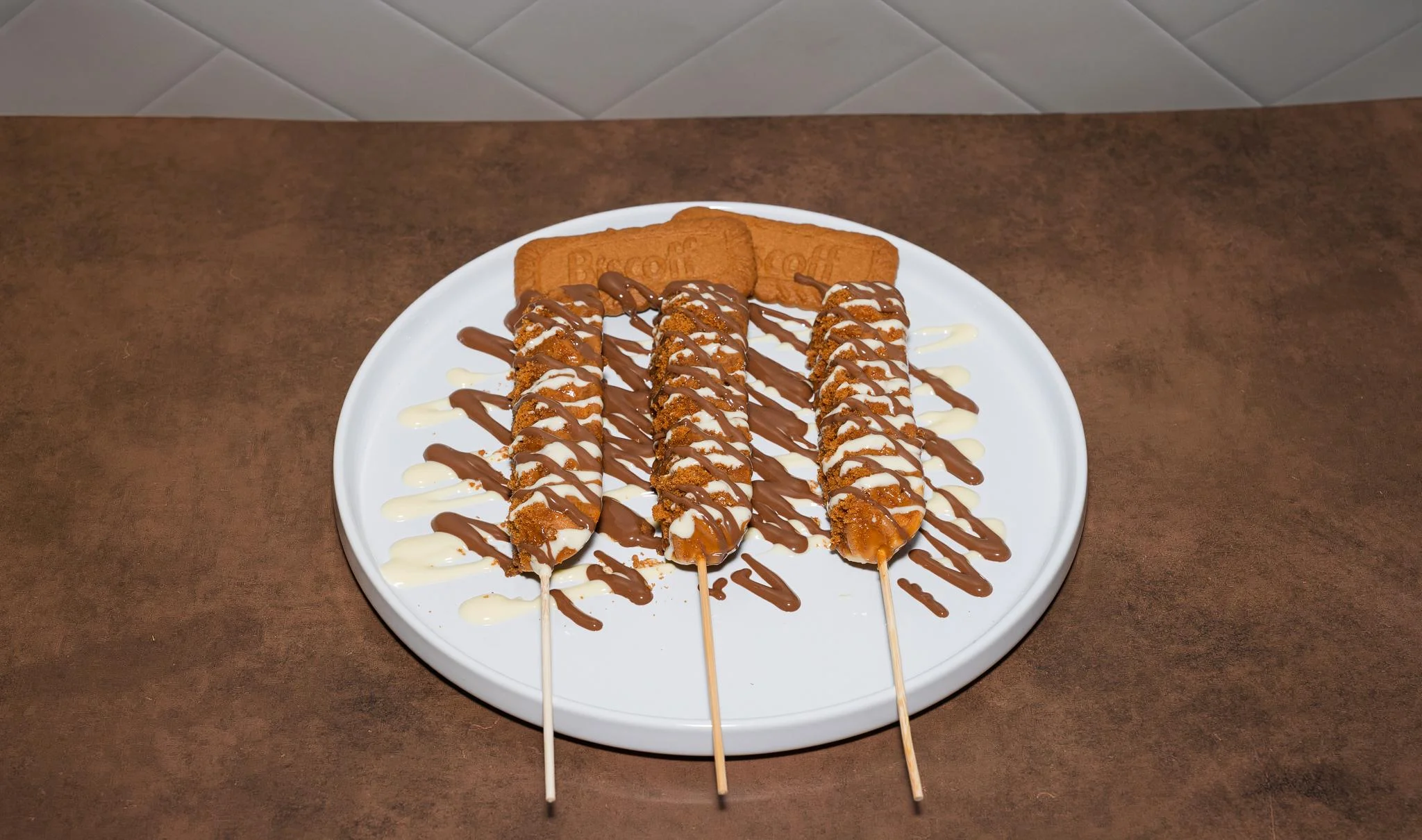 Three dessert skewers drizzled with chocolate and decorated with cookie pieces, arranged on a white plate.