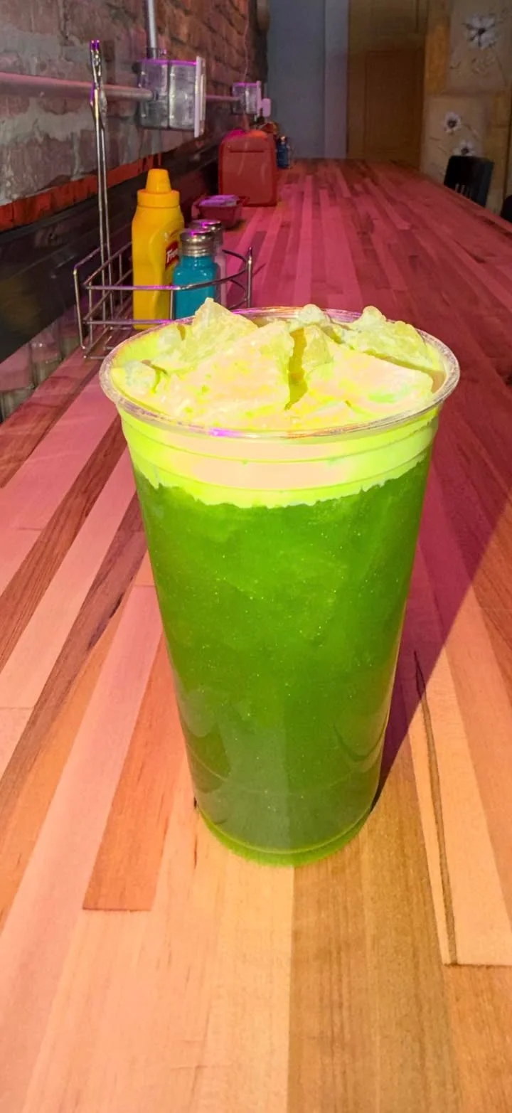 A vibrant green drink served in a clear cup, filled with ice and topped with a light foam. The drink sits on a wooden counter.