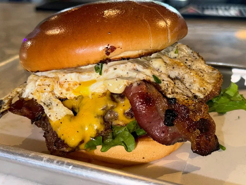 A delicious burger featuring a toasted bun, layers of bacon, cheese, lettuce, and a fried egg. The burger is presented on a tray, showcasing its hearty ingredients.