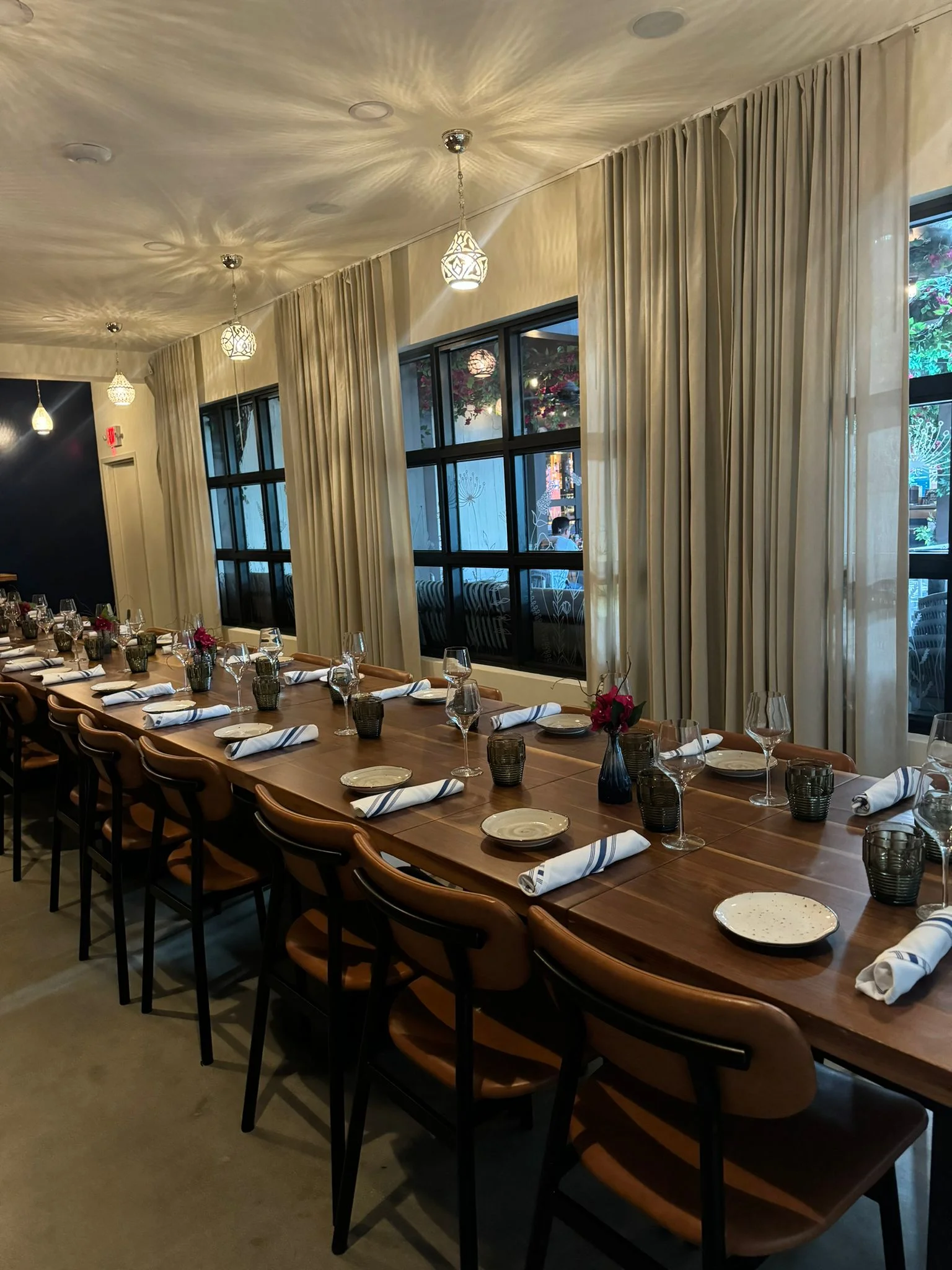 Elegant dining setup with a long wooden table and brown chairs. Table is set with white napkins, plates, wine glasses, and small vases with red flowers. Cream curtains and warm pendant lights add a sophisticated ambiance.