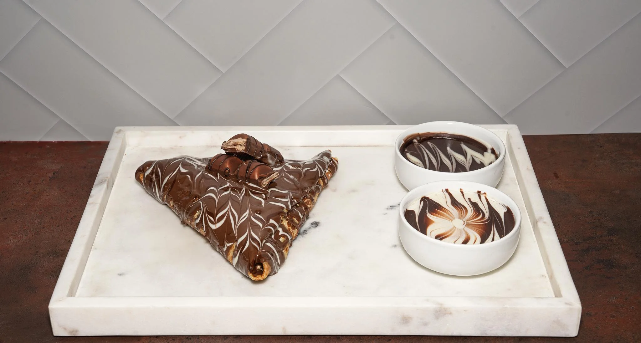 A triangular chocolate confection placed next to two bowls of swirled chocolate, all presented on a marble tray.