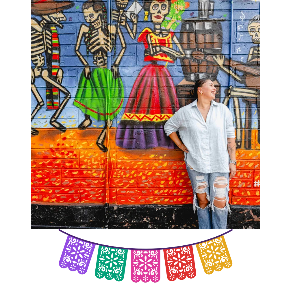 Sandra Lopez, owner of Friducha in Brooklyn, NY, smiling in front of a colorful Day of the Dead mural.