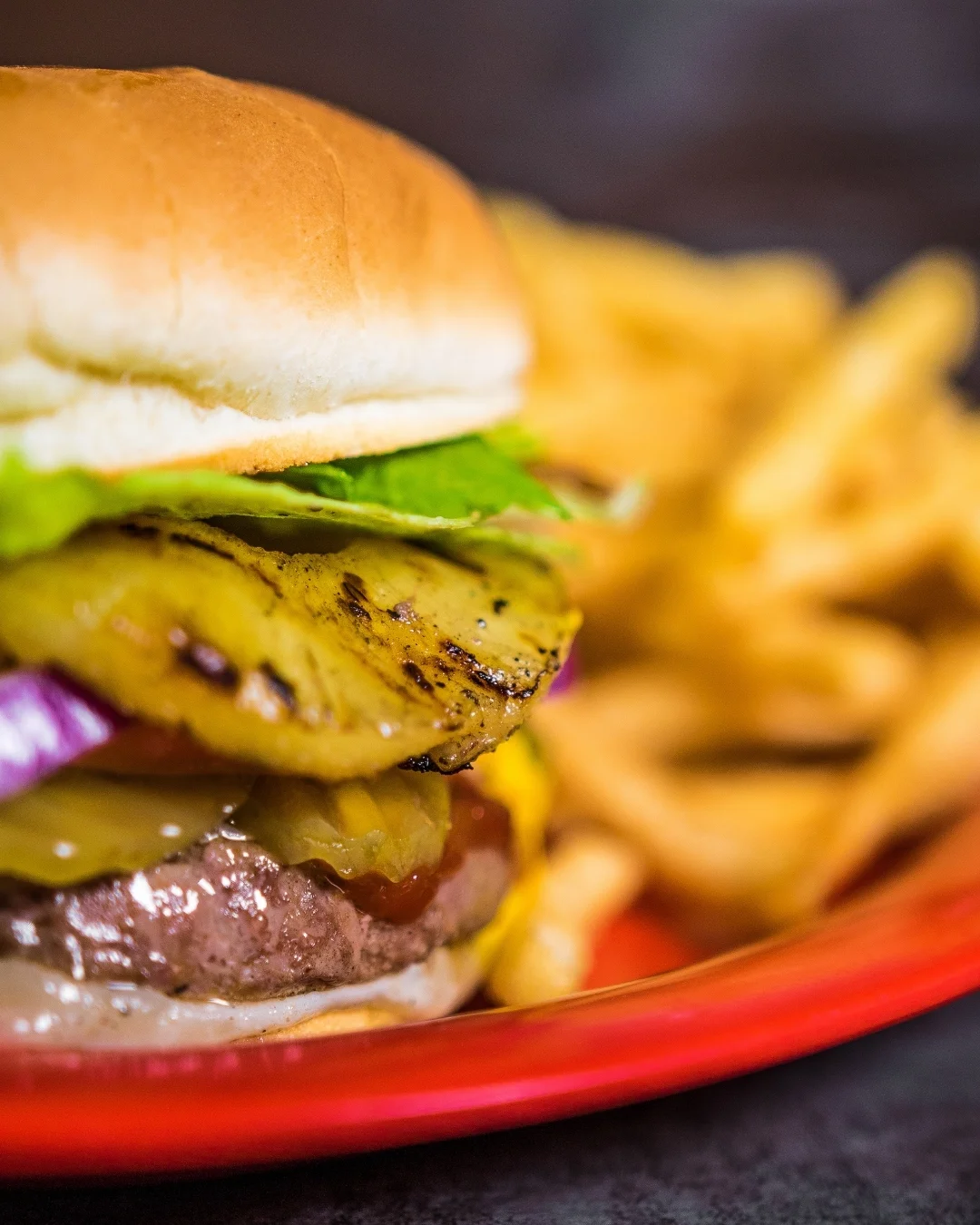 A juicy hamburger stacked with two patties, melted cheese, fresh lettuce, tomato, and grilled pineapple, served alongside a portion of sweet potato fries.