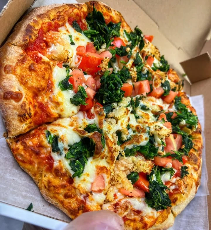An assortment of toppings on a pizza including fresh tomatoes and leafy greens. The pizza is placed inside a box.