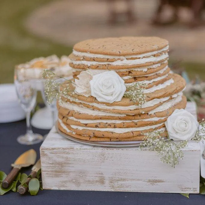 A tiered cookie cake made of large stacked cookies with frosting layers in between, decorated with white roses and baby’s breath.