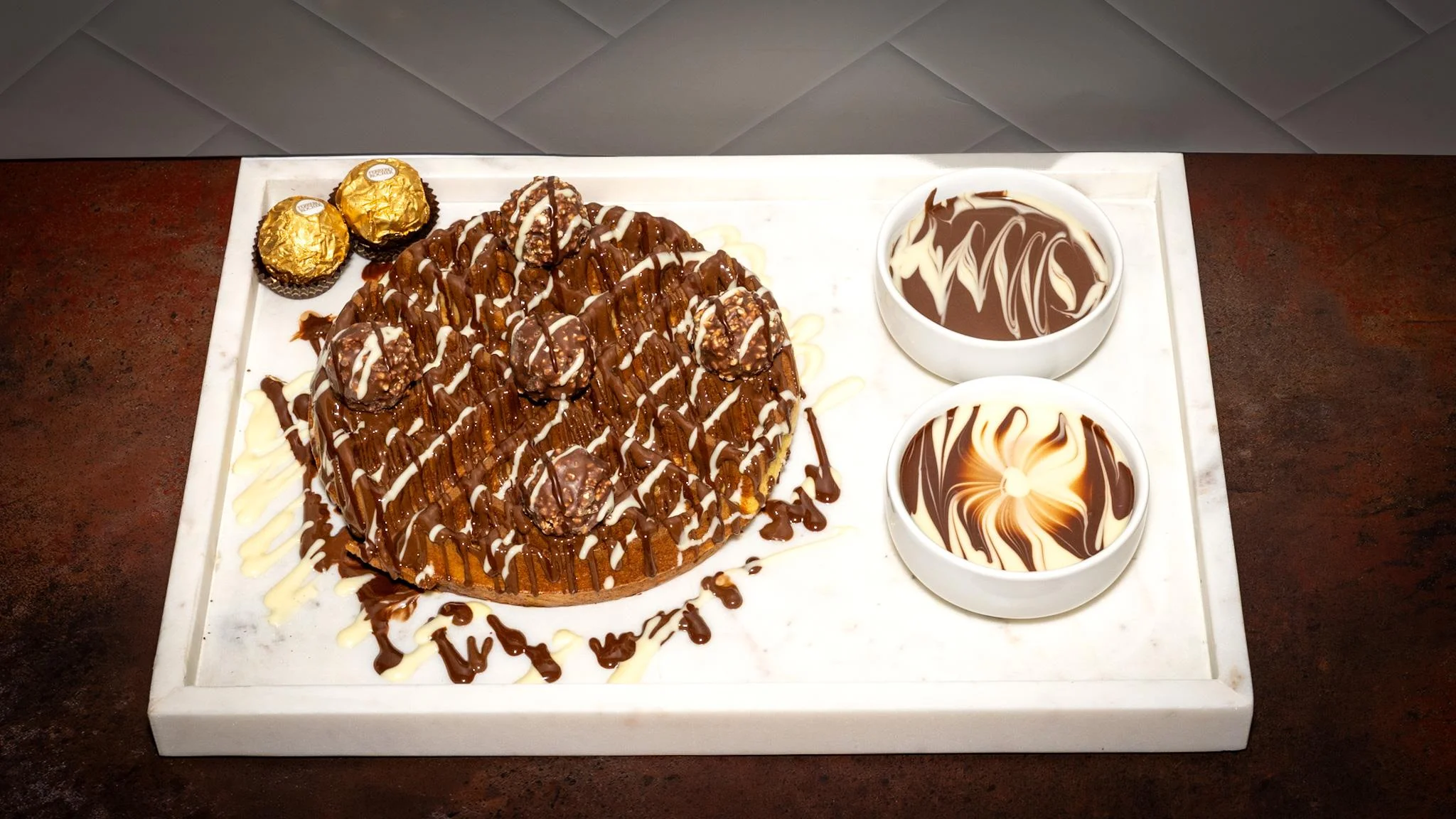 A round cake decorated with chocolate and golden hazelnut treats, accompanied by two bowls of swirled chocolate sauce.