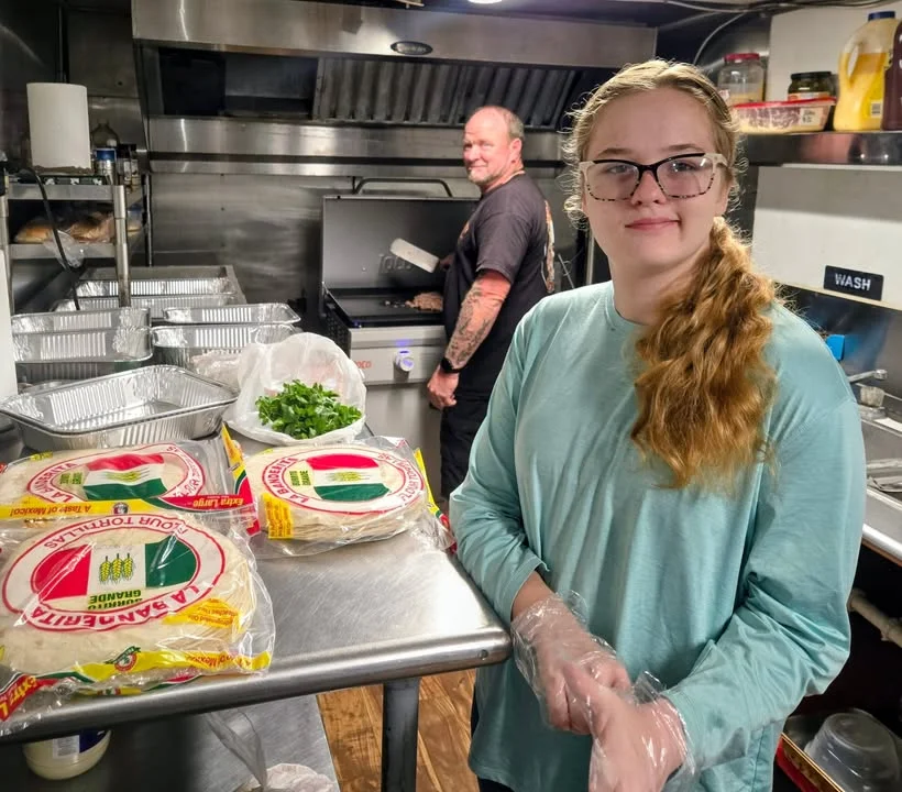 A young girl and an adult preparing food in a kitchen, with tortillas and fresh ingredients visible on the counter.