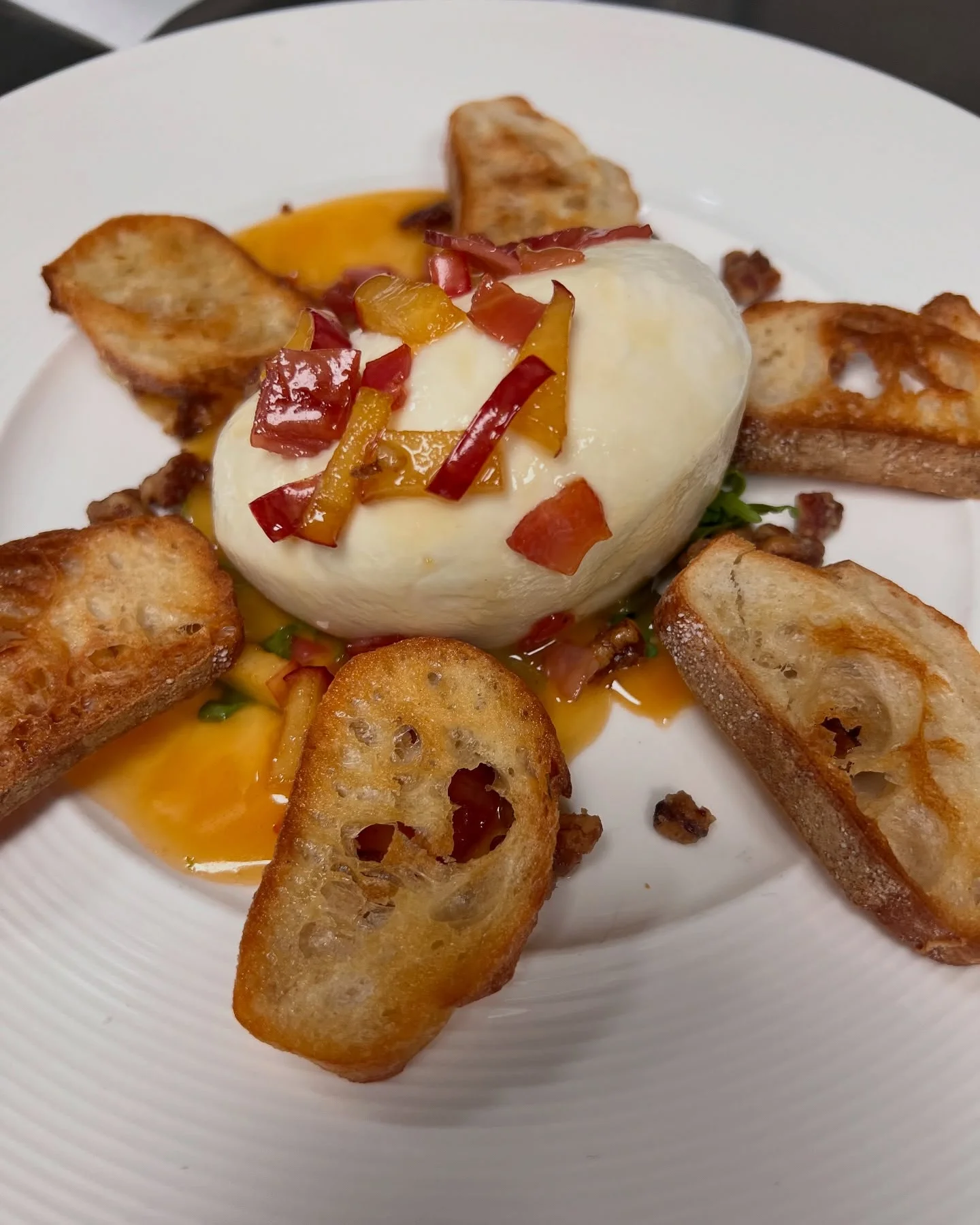 Plated burrata cheese topped with diced red and yellow peppers, surrounded by toasted crostini slices and drizzled with a vibrant orange sauce. Garnished with crumbled bits and served on a white dish with a modern, elegant presentation