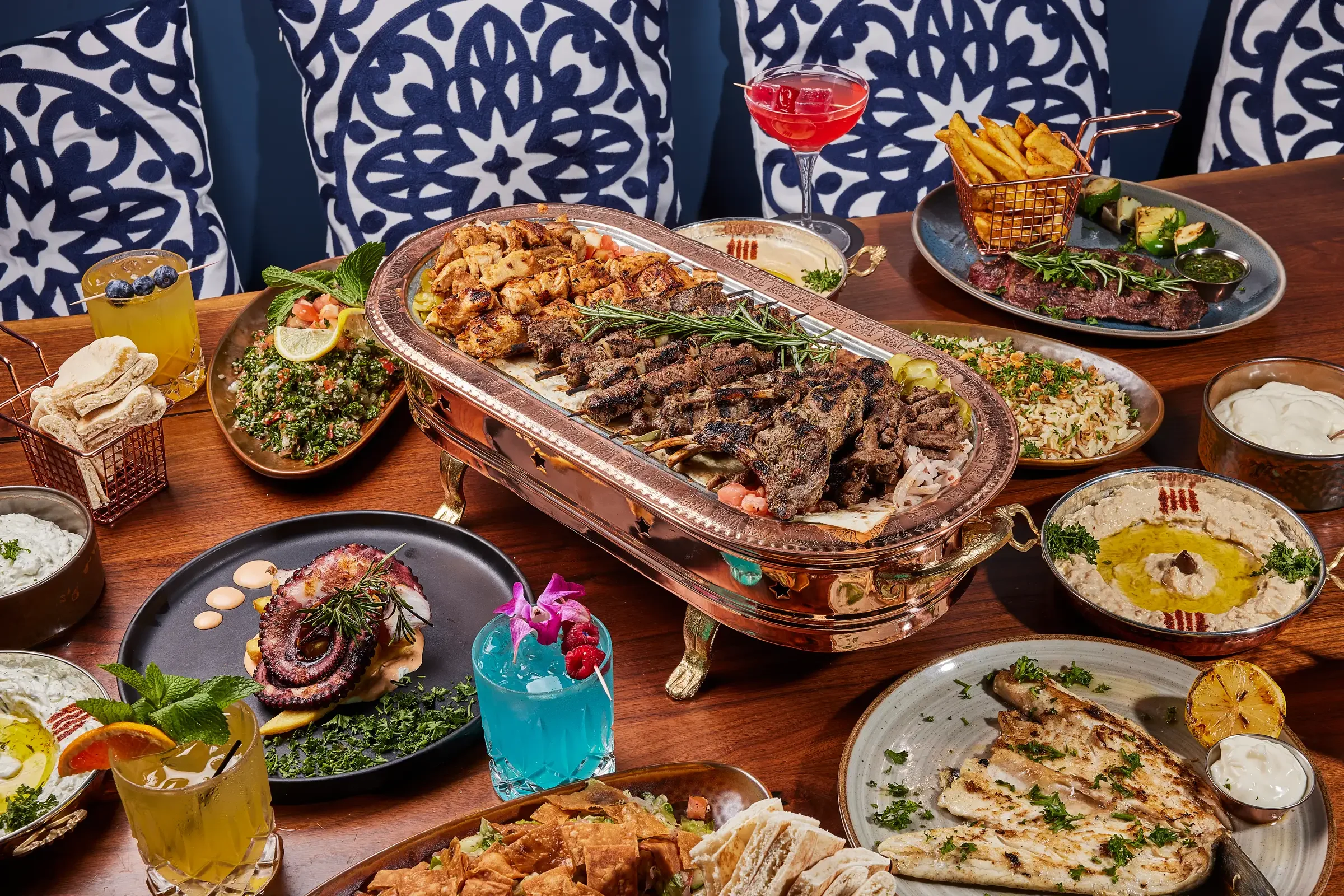 A vibrant array of Mediterranean dishes on a wooden table, featuring grilled meats on a large platter, colorful drinks, hummus, and dips, with decorative blue-patterned cushions in the background.