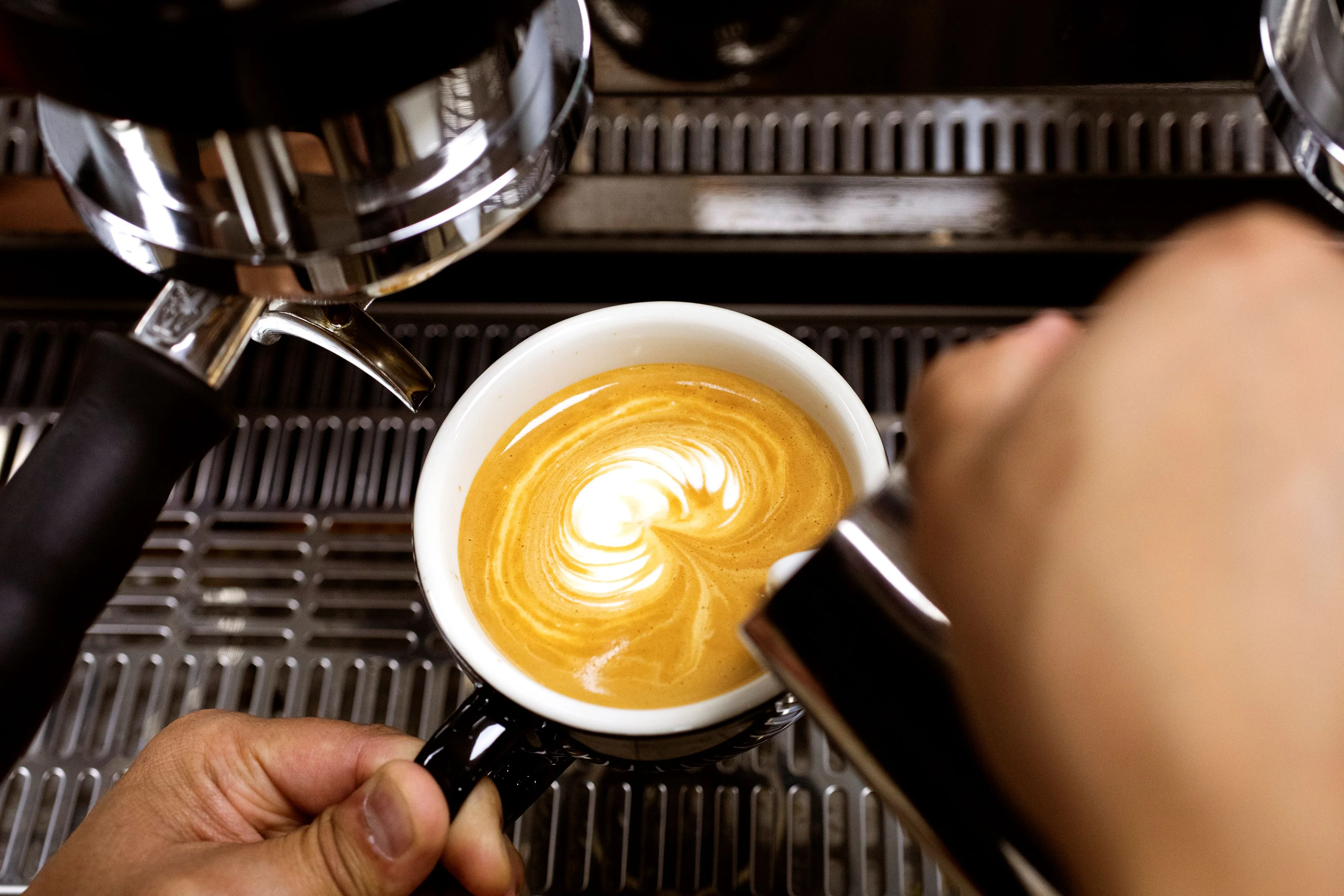 A barista crafts latte art in a white cup beneath a portafilter. The golden espresso swirls blend with creamy milk, creating a serene heart pattern.